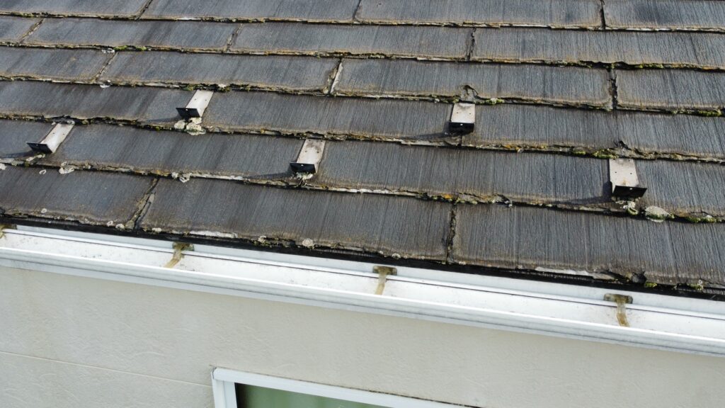 カバー工事前のスレート屋根の写真です。スレート屋根は、屋根材自体に防水機能がないため、表面の塗膜によって防水性を保っています。﻿紫外線で塗膜が劣化すると色褪せ、コケや藻の発生、ひび割れ、反り、欠けなどが症状として表れます。