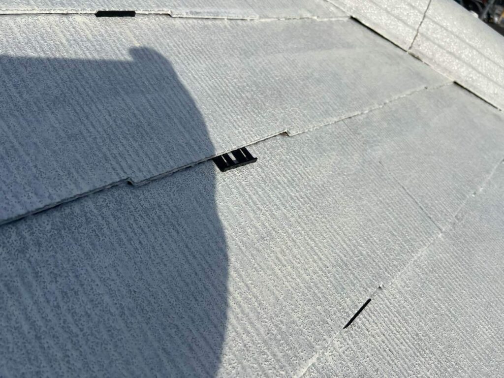 タスペーサーを挿入する様子です。タスペーサーにはシングル工法とダブル工法があり、ユウマペイントではダブル工法を採用しています。タスペーサーダブル工法とは、スレート屋根塗装で使われる「縁切り」という工程の一つで、1枚の約90cm幅のスレート屋根材に対して、両端から15cm内側にタスペーサーを2個（左右それぞれ1個ずつ）挿入する方法です。これはタスペーサーの開発元が推奨する工法で、下塗り乾燥後に差し込み、塗料でスレートの隙間が塞がってしまうのを防ぎ、雨漏りや瓦の割れを防ぐ目的があります。90cm幅の屋根材は、塗装で隙間が塞がりやすいため、より確実に縁切り（隙間を確保）するために2個挿入するダブル工法が推奨されています。