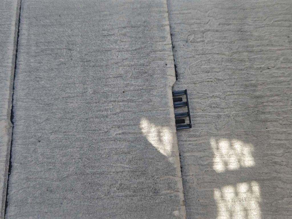 タスペーサーを挿入する様子です。タスペーサーにはシングル工法とダブル工法があり、ユウマペイントではダブル工法を採用しています。タスペーサーダブル工法とは、スレート屋根塗装で使われる「縁切り」という工程の一つで、1枚の約90cm幅のスレート屋根材に対して、両端から15cm内側にタスペーサーを2個（左右それぞれ1個ずつ）挿入する方法です。これはタスペーサーの開発元が推奨する工法で、下塗り乾燥後に差し込み、塗料でスレートの隙間が塞がってしまうのを防ぎ、雨漏りや瓦の割れを防ぐ目的があります。90cm幅の屋根材は、塗装で隙間が塞がりやすいため、より確実に縁切り（隙間を確保）するために2個挿入するダブル工法が推奨されています。