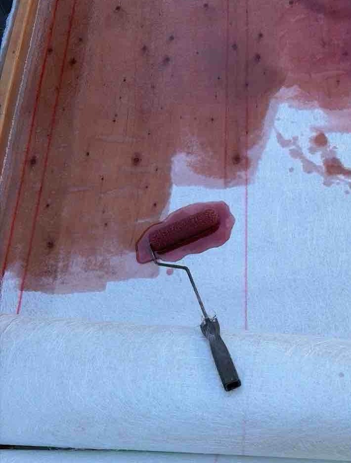 敷き詰めた「ガラスマット（白いシート状の補強材）」の上に、防水性のある「不飽和ポリエステル樹脂（茶褐色の液体）」を専用ローラーで塗布しています。単に樹脂を塗るだけでなく、ローラーでしっかりと押さえつけることで、ガラスマットの繊維の奥まで樹脂をしみ込ませ（含浸）、同時に内部の気泡を押し出します（脱泡）。この作業を丁寧に重ねることで、軽量ながら極めて強靭で、継ぎ目のない完全な水密性を持った防水層が形成されます。