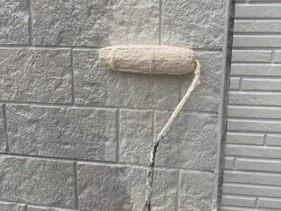 外壁の下塗り工程の様子です。ユウマペイントでは、塗料の耐久性を確実なものにするため、サイディング外壁の下塗りを2回に分けて丁寧に行うこだわりの仕様を採用しています。1回目の下塗りでは「日本ペイント ファイン浸透シーラー」を塗布し、傷んだサイディング材にしっかりと浸透させて下地を強固に固め、塗料の密着性を高めるための土台を作ります。続いて2回目の下塗りとして「日本ペイント 水性シリコンエポサーフ」を重ねます。この塗料は高い柔軟性を持っているため、外壁に発生した微細なひび割れ（ヘアクラック）をしっかりと埋めるとともに、高い密着力で下地自体の劣化を防ぎ、この後に重ねる上塗り塗料の性能を最大限に引き出す効果があります。写真のように、元のレンガ調の色がしっかりと隠れるまで真っ白に塗りつぶして完璧な下地を作ることで、上塗りの仕上がりと持ちが格段に良くなります。