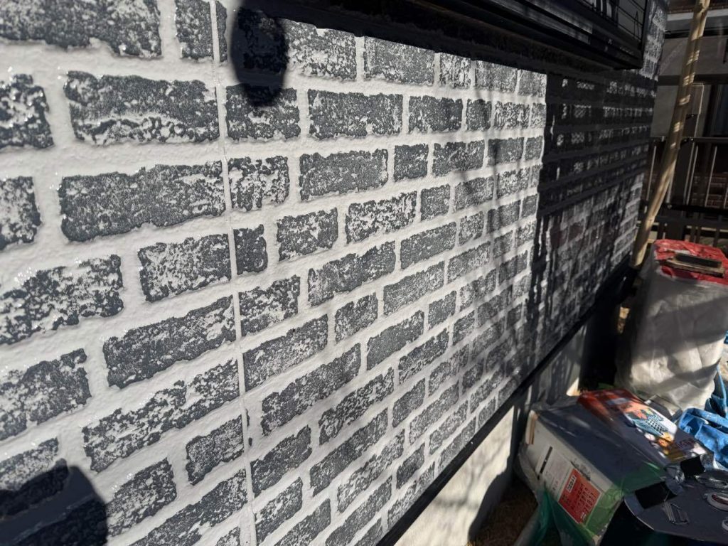 階部分のタイル調サイディングに施した、グラデーション塗装（2色塗り）の工程です。通常の単色塗りでは目地も表面も同じ色で塗りつぶされ、元のレンガ調の立体感や風合いが完全に失われてしまいます。そこで、まずは目地の色となるベースカラーで全体を塗り上げ、乾燥後にユウマペイント独自の特殊な施工方法を用いて、出っ張っている表面部分にだけアクセントカラー（ダークグレー系）を丁寧に重ねていきます。職人の緻密な手作業によってこのひと手間を加えることで、目地と表面で綺麗に色が分かれ、立体的で重厚感のある美しい意匠性をよみがえらせることができます。