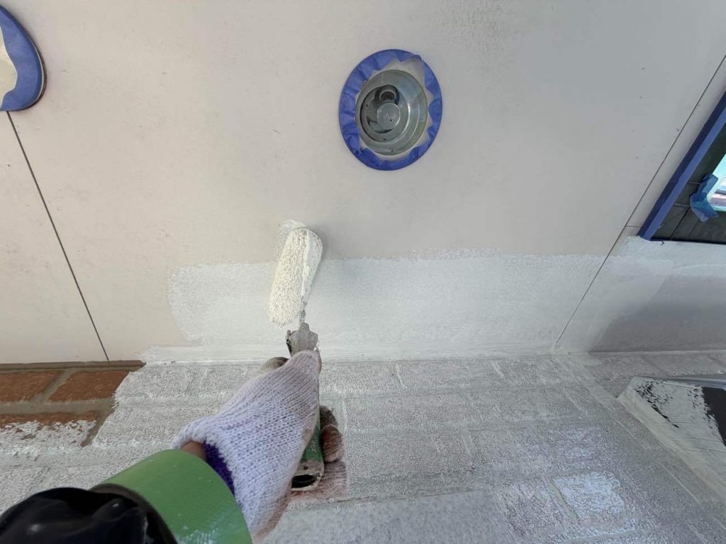 軒天井（のきてん）の下塗り（シーラー塗布）を行っている様子です。屋根の裏側にあたる軒天井は直接雨水は当たらないものの、日陰になりやすく湿気が滞留するため、カビや傷みが発生しやすい環境にあります。そのため、まずは専用の下塗り材「シーラー」をローラーでたっぷりと塗布し、下地の奥深くまでしっかりと浸透させます。これにより、素材の表面を強固に固めて塗料の無駄な吸い込みを抑えるとともに、この後に塗布する「透湿性防カビ塗料（上塗り）」をピタッと密着させるための強力な接着剤の役割を果たしてくれます。塗膜の早期剥がれを防ぎ、美しい仕上がりを長期にわたって長持ちさせるためには決して欠かすことのできない大切な基礎工程です。また、写真に写っている照明や換気口などの設備は、塗料が付着しないように事前にテープ等でしっかりと養生（保護）してから丁寧な作業を進めています。