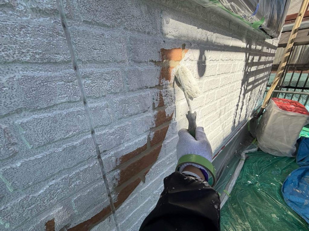 外壁の下塗り工程の様子です。ユウマペイントでは、塗料の耐久性を確実なものにするため、サイディング外壁の下塗りを2回に分けて丁寧に行うこだわりの仕様を採用しています。1回目の下塗りでは「日本ペイント ファイン浸透シーラー」を塗布し、傷んだサイディング材にしっかりと浸透させて下地を強固に固め、塗料の密着性を高めるための土台を作ります。続いて2回目の下塗りとして「日本ペイント 水性シリコンエポサーフ」を重ねます。この塗料は高い柔軟性を持っているため、外壁に発生した微細なひび割れ（ヘアクラック）をしっかりと埋めるとともに、高い密着力で下地自体の劣化を防ぎ、この後に重ねる上塗り塗料の性能を最大限に引き出す効果があります。写真のように、元のレンガ調の色がしっかりと隠れるまで真っ白に塗りつぶして完璧な下地を作ることで、上塗りの仕上がりと持ちが格段に良くなります。