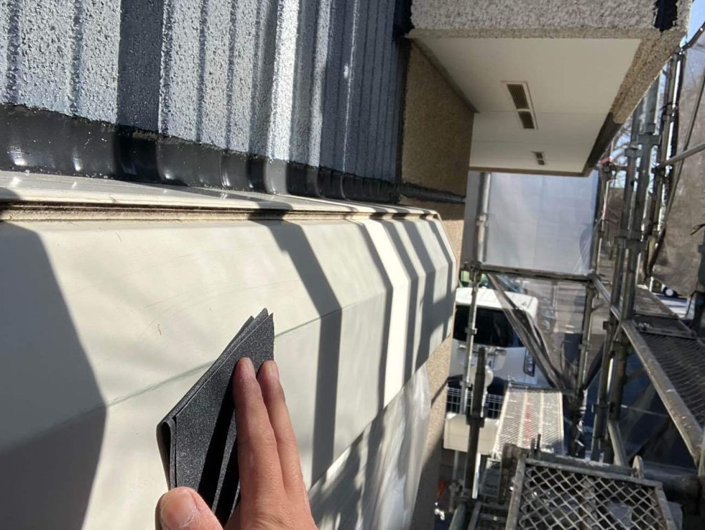窓上部に設置されている金属製のシャッターボックス、下地処理の工程です。太陽光が直接当たるため塗膜の劣化が進行しやすい箇所です。サンドペーパーなどを使い、表面の旧塗膜やサビ、汚れを研磨して落とすケレン作業を行います。この作業には、表面を平滑に整えると同時に、微細な傷をつけて塗料の密着を高める「目荒らし」の効果もあり、塗装の耐久性を左右する重要な工程となります。