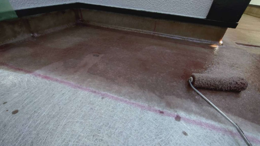FRP防水工事の施工中の様子で下地処理とプライマー塗布を終えた床面に強度を高めるためのガラスマットを敷き詰めその上から防水層の主体となるポリエステル樹脂をローラーでたっぷりと含ませて空気を抜きながら密着させている工程です。